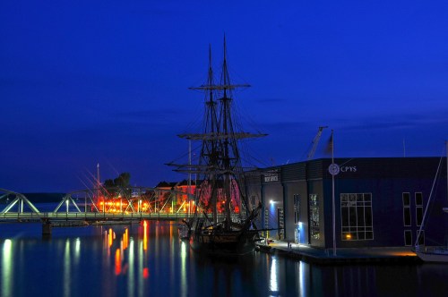 A tall ship docked by a modern building, illuminated by colorful lights, reflecting in calm waters at dusk.