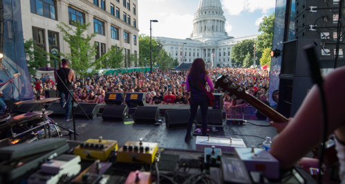 A live band performs on stage in front of a large crowd, with a historic building in the background.