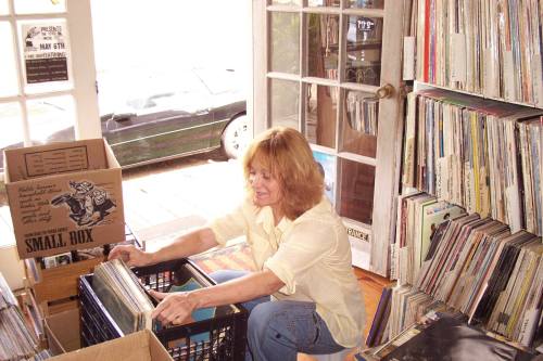 A woman with wavy hair organizes vinyl records in a cozy room filled with boxes and shelves of albums.