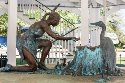 A bronze statue of a woman kneeling beside a heron, set in a park under a gazebo.