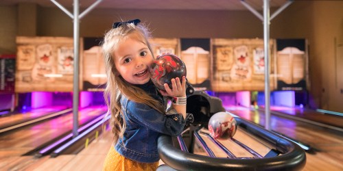 A smiling girl holds a bowling ball while standing at a bowling lane, ready to play.