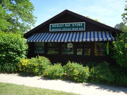 A quaint store with a striped awning, surrounded by greenery and flowers, displaying a sign for groceries and snacks.