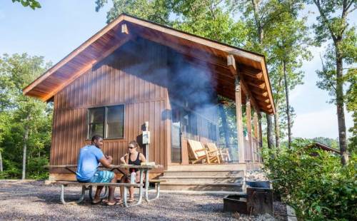 A couple sits at a picnic table outside a wooden cabin surrounded by trees, with smoke rising from a nearby fire pit.