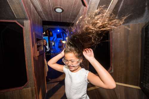 A girl with long hair spins joyfully in a dimly lit, wooden-themed space, wearing glasses and enjoying the moment.