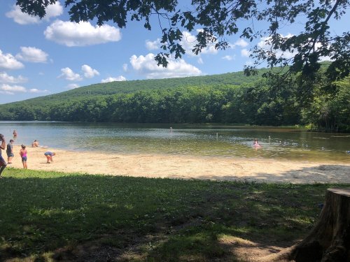 A serene lake surrounded by green hills, with children playing on the sandy shore under a blue sky with fluffy clouds.