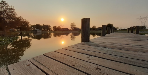 A serene sunset over a calm lake, reflecting the sun and trees, with a wooden dock in the foreground.