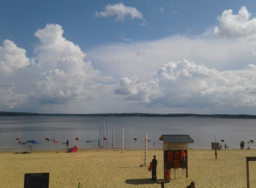 A sandy beach with people, lifebuoys, and a calm lake under a cloudy sky.