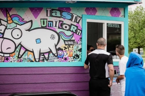 A colorful food truck named "Tasty Unicorn" with a whimsical unicorn mural, customers placing orders at the window.