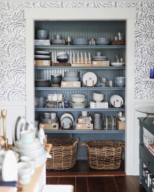 A neatly organized shelf displaying various dishes, glasses, and woven baskets against a patterned wallpaper backdrop.