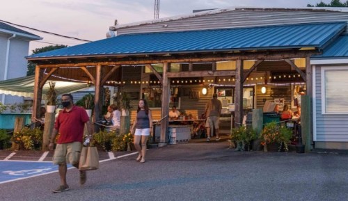 A casual outdoor dining area with people inside and outside, featuring string lights and a blue roof at dusk.