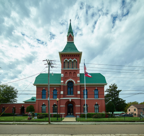 Historic red brick building with a green roof and tall spire, surrounded by trees and power lines under a cloudy sky.