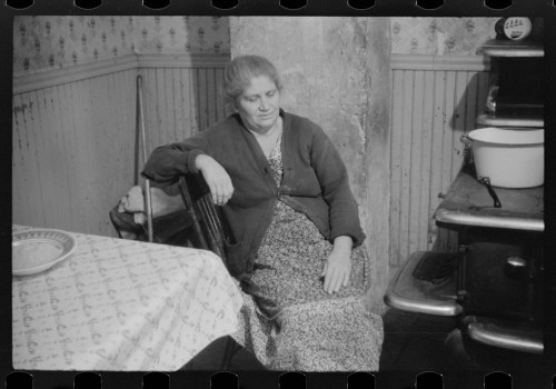 A woman in a long dress sits in a chair beside a table in a vintage kitchen, looking contemplative.