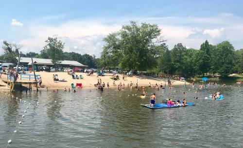 A sunny beach scene with people swimming, lounging, and enjoying a lake surrounded by trees and a sandy shore.