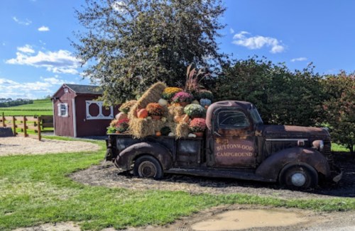 An old truck filled with colorful autumn decorations parked near a red barn and green fields under a blue sky.