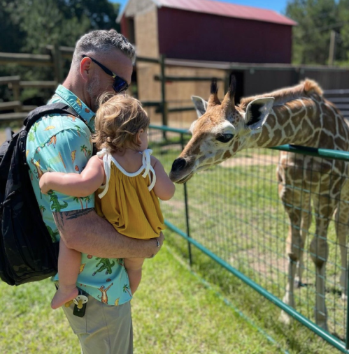 A man holds a toddler as they interact with a giraffe at a petting zoo on a sunny day.