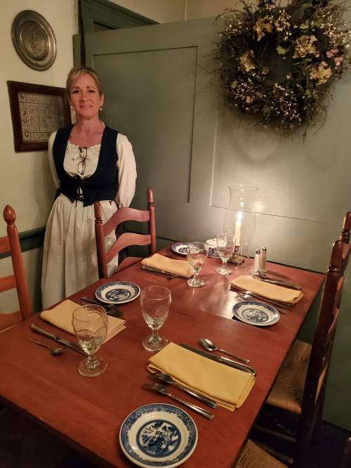 A woman in period attire stands beside a candlelit dining table set with glassware and plates in a cozy room.
