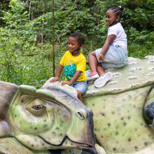 Two children play on a large, colorful dinosaur sculpture in a lush green park.
