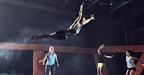 A person jumps mid-air at a trampoline park, surrounded by three others, with a dramatic lighting effect.