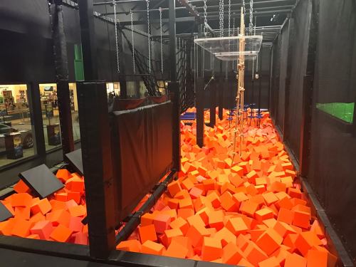 A trampoline park featuring a large pit filled with bright orange foam cubes and safety nets surrounding the area.