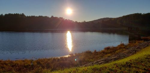 A serene lake at sunset, with the sun reflecting on the water and surrounded by trees and hills.
