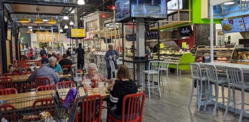 A bustling food hall with diners at tables, food counters, and screens displaying menus and entertainment.