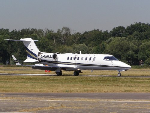 A white and blue private jet taxiing on a runway with trees in the background.