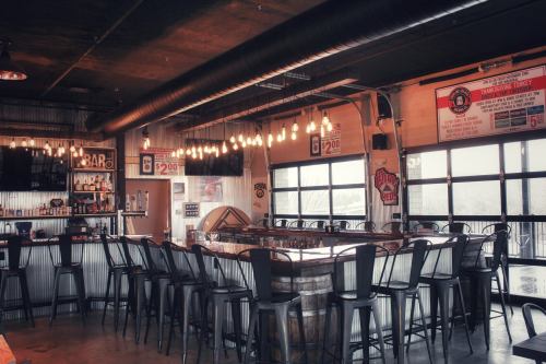 A modern bar interior with high stools, a wooden counter, and warm lighting, featuring large windows and a rustic vibe.