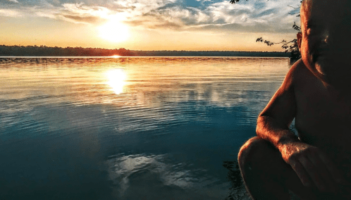 A serene sunset over a calm lake, with a person sitting at the water's edge, reflecting on the peaceful scene.