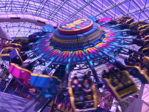 A colorful amusement park ride spins under a glass roof, with riders enjoying the thrill.