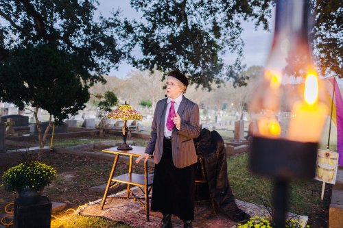 A person in formal attire speaks at a cemetery, with a lamp and table nearby, surrounded by gravestones and trees.