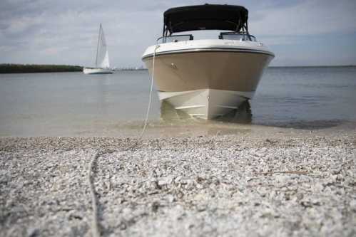 A boat on a sandy shore with calm water, another sailboat in the background under a cloudy sky.