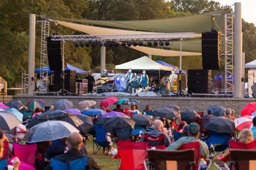 A crowd with umbrellas watches a live band perform on an outdoor stage during a concert in a park.
