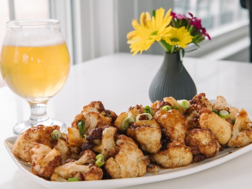 A platter of crispy cauliflower bites drizzled with sauce, garnished with green onions, alongside a glass of beer and flowers.