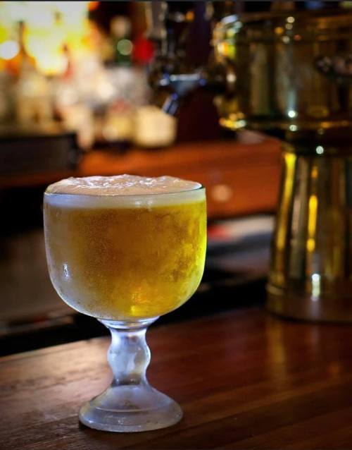 A frosty glass of beer sits on a wooden bar, with a blurred background of bottles and a beer tap.