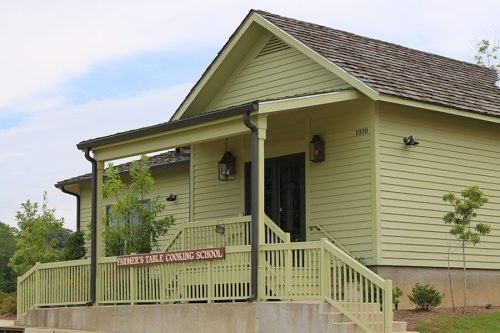 A green building with a porch and sign reading "Farmer's Table Cooking School" surrounded by trees and blue sky.