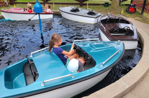 A girl steers a small blue boat in a pond, with other boats and children playing in the background.