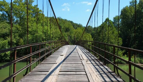 A wooden suspension bridge stretches over a river, surrounded by lush green trees under a clear blue sky.