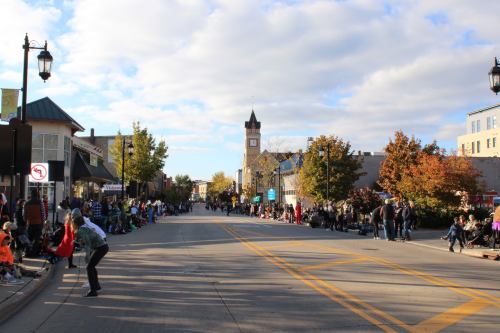 A wide street lined with spectators waiting for a parade, featuring trees and a clock tower in the background.