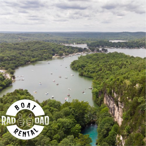 Aerial view of a river surrounded by greenery, with boats scattered on the water and a cliff in the foreground.