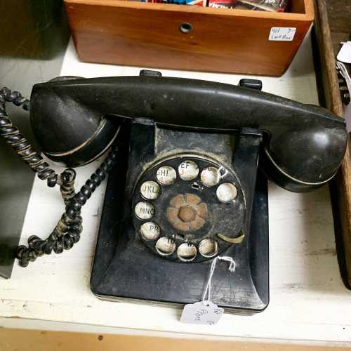 A vintage rotary phone with a black body and a coiled cord, placed on a table among other items.