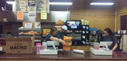 Three employees work behind a concession stand, preparing snacks and drinks in a casual setting.