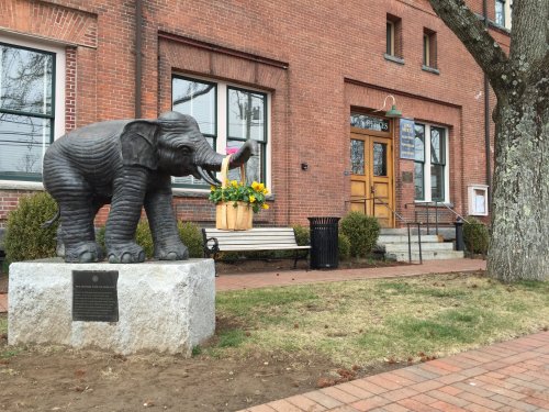 A bronze elephant statue holding a basket of flowers in front of a brick building with a bench and tree nearby.