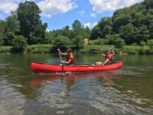 Two people paddle a red canoe on a calm river surrounded by lush green trees under a blue sky.