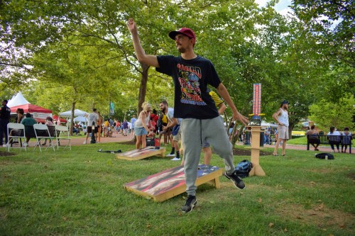 A man in a cap throws a bean bag at a cornhole board in a park filled with people and trees.