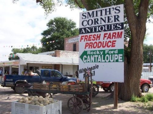 Sign for Smith's Corner Antiques featuring fresh farm produce, including Rocky Ford cantaloupe, with a cart of melons nearby.