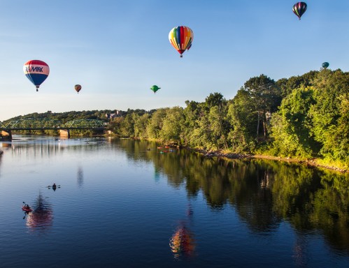 Colorful hot air balloons float above a calm river, reflecting in the water, surrounded by lush greenery and a bridge.