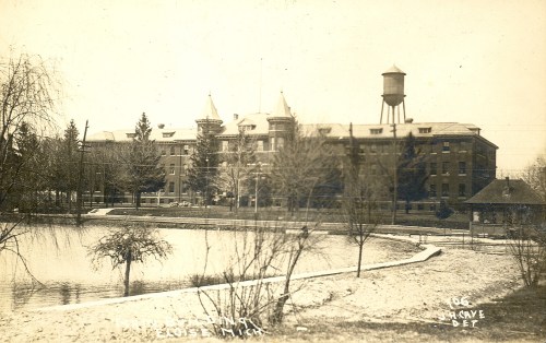 Historic building with towers beside a pond, surrounded by trees, in a sepia-toned photograph.