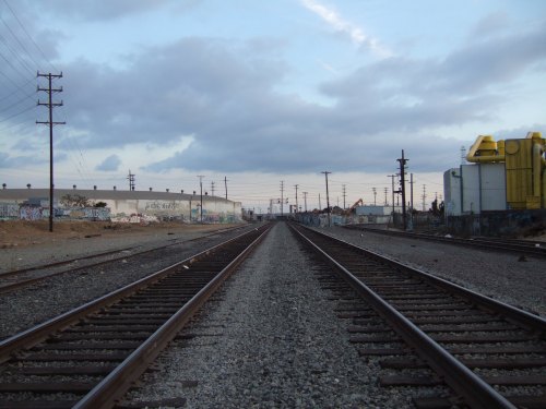 A view of railway tracks stretching into the distance, flanked by industrial buildings and a cloudy sky.