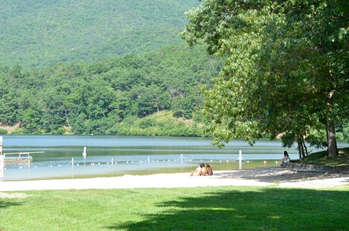 A serene lakeside scene with people sitting on the shore, surrounded by lush greenery and mountains in the background.