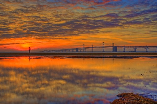 A vibrant sunset reflects on water with a bridge silhouette in the background and a person standing nearby.